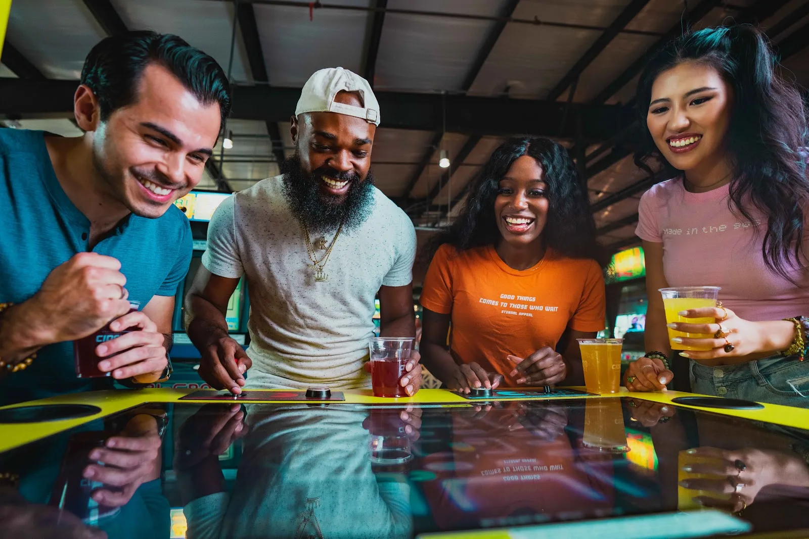 Four friends holding drinks and laughing over a tabletop arcade game at Cidercade Houston