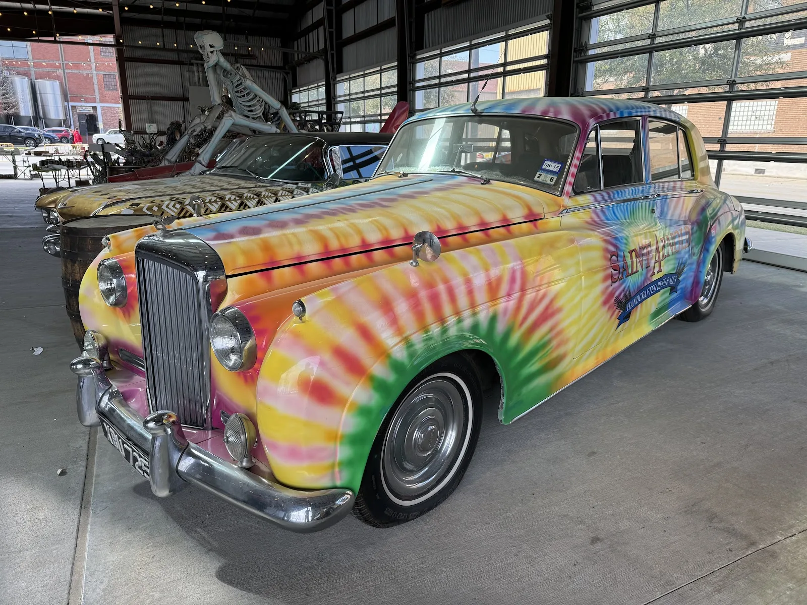 A tie-dye painted vintage car with the Saint Arnold logo parked under cover at the Saint Arnold brewery
