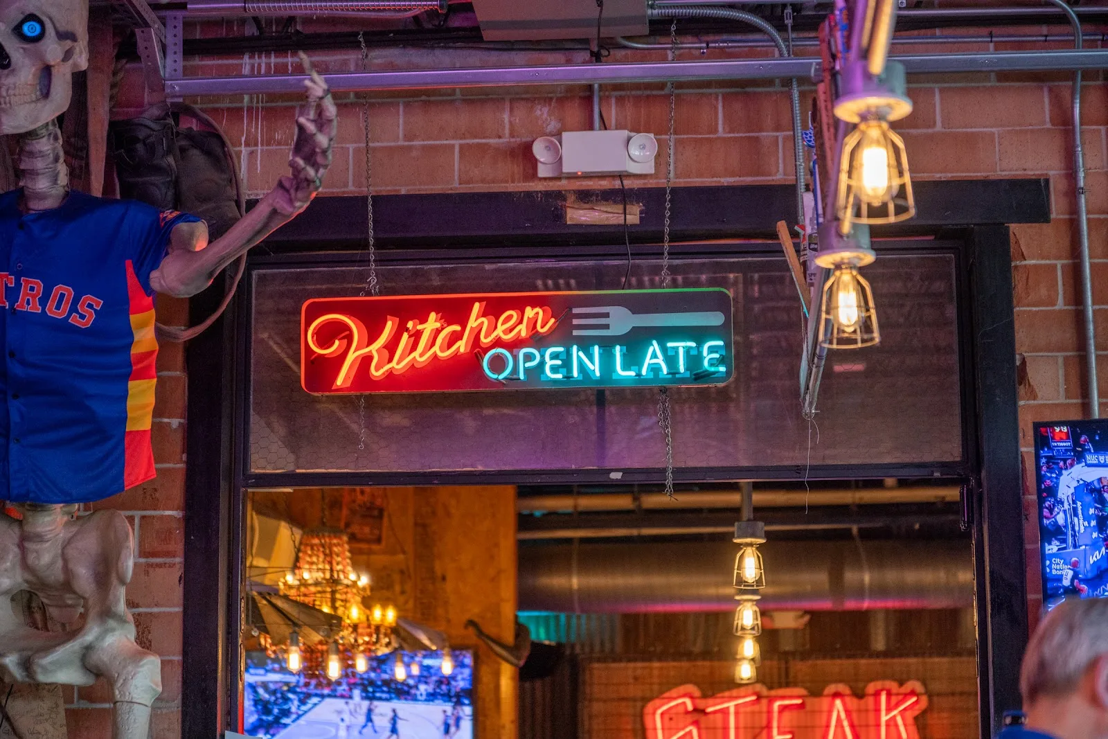 A neon Kitchen Open Late sign beside a skeleton in an Astros jersey at Truck Yard in EaDo