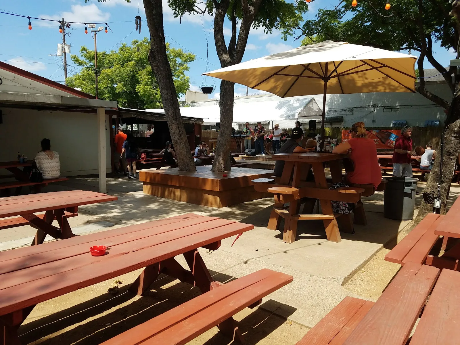 Red picnic tables under trees and string lights at the West Alabama Ice House with a band playing in the background